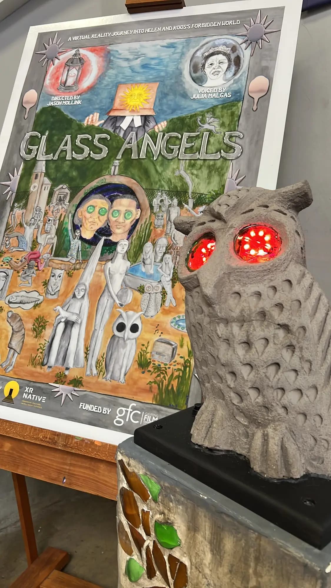 The painted Glass Angels poster on an easel, beside an owl figure whose eyes glow red