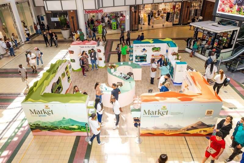 A busy indoor market at the V&A Waterfront. Orange and white stalls read Market and Made in the Cape. Shoppers stand at the counters. A Havaianas store is behind them.