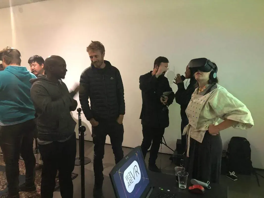 A WeAReVR Jozi meetup, May 2019. A guest stands in a VR headset with one hand on their hip while the people around them laugh and talk. A rope-barrier post and a laptop showing the WE ARE VR logo are in the foreground.