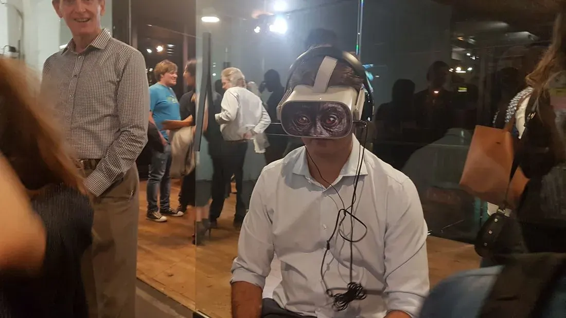 A WeAReVR Jozi meetup, October 2019. A visitor sits in a VR headset with its cables hanging down, while someone stands beside them smiling and other guests talk in the background.