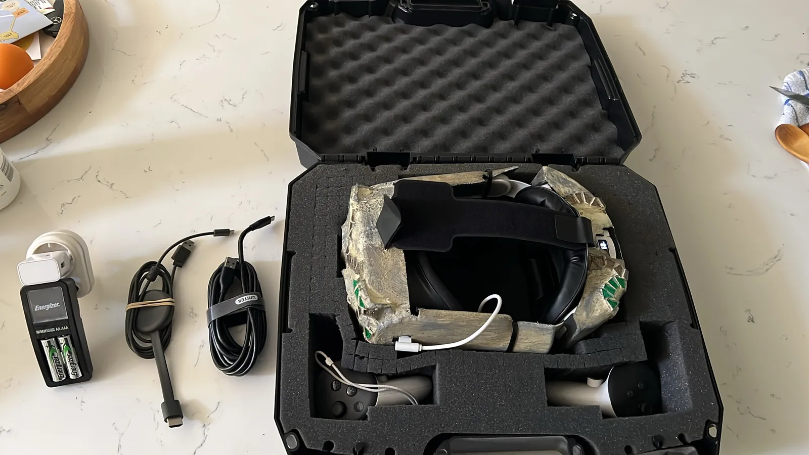 The Glass Angels headset in its mosaic shell, packed into a foam-lined hard case with two controllers. A battery charger and two cables lie on the counter beside it.