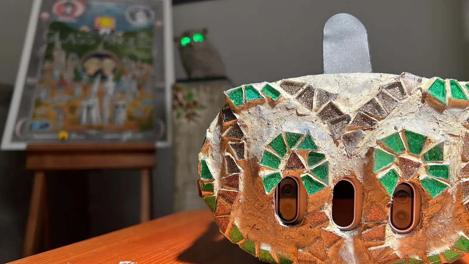 Close-up of the mosaic headset shell, green glass rings around the camera openings like an owl's eyes. The poster and the owl sculpture sit out of focus behind it.