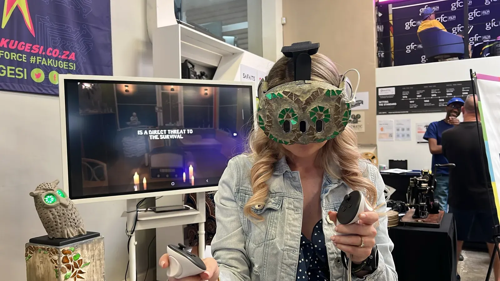 A visitor wearing the mosaic owl headset holds two Quest controllers at the Glass Angels stand at Fak'ugesi. Behind them, a screen mirrors the view inside the headset, with candles and the words "is a direct threat to the survival". An owl sculpture with glowing green eyes sits on a mosaic plinth to the left, and other festival-goers stand around in the background.