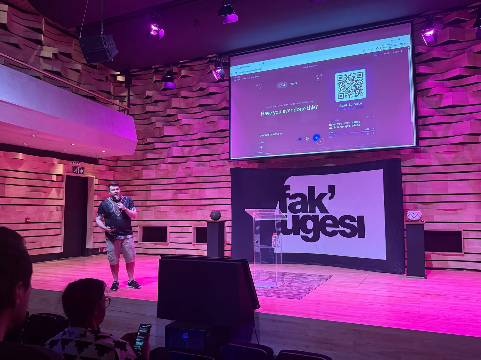 Deac on stage at Fak'ugesi under pink lights, mic in hand. The big screen shows the opening question, "Have you ever done this?", with a QR code to vote. In the front row, someone is holding a phone.